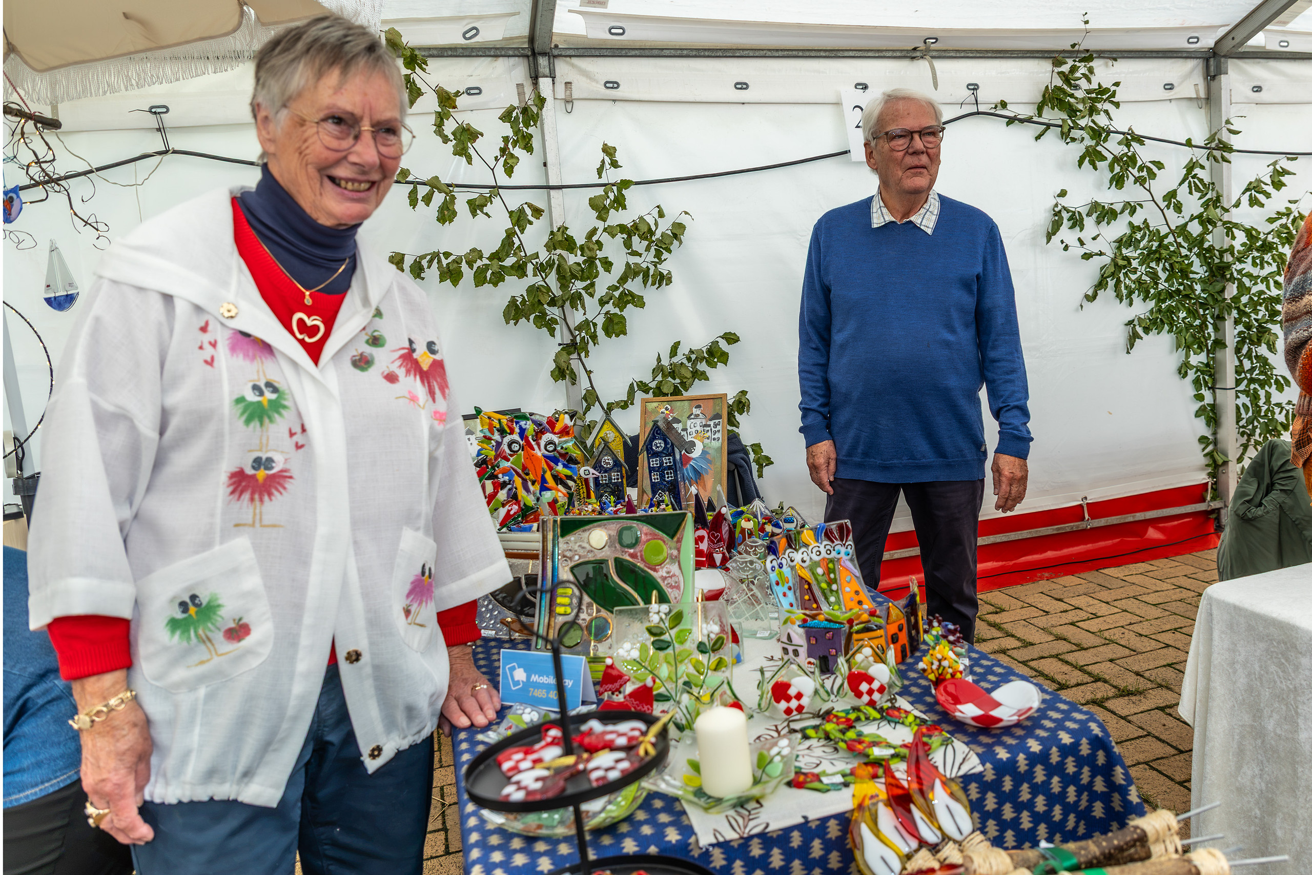 Smilende kvinde i broderet jakke ved en farverig bod i festivalteltet på Gråsten Æblefestival