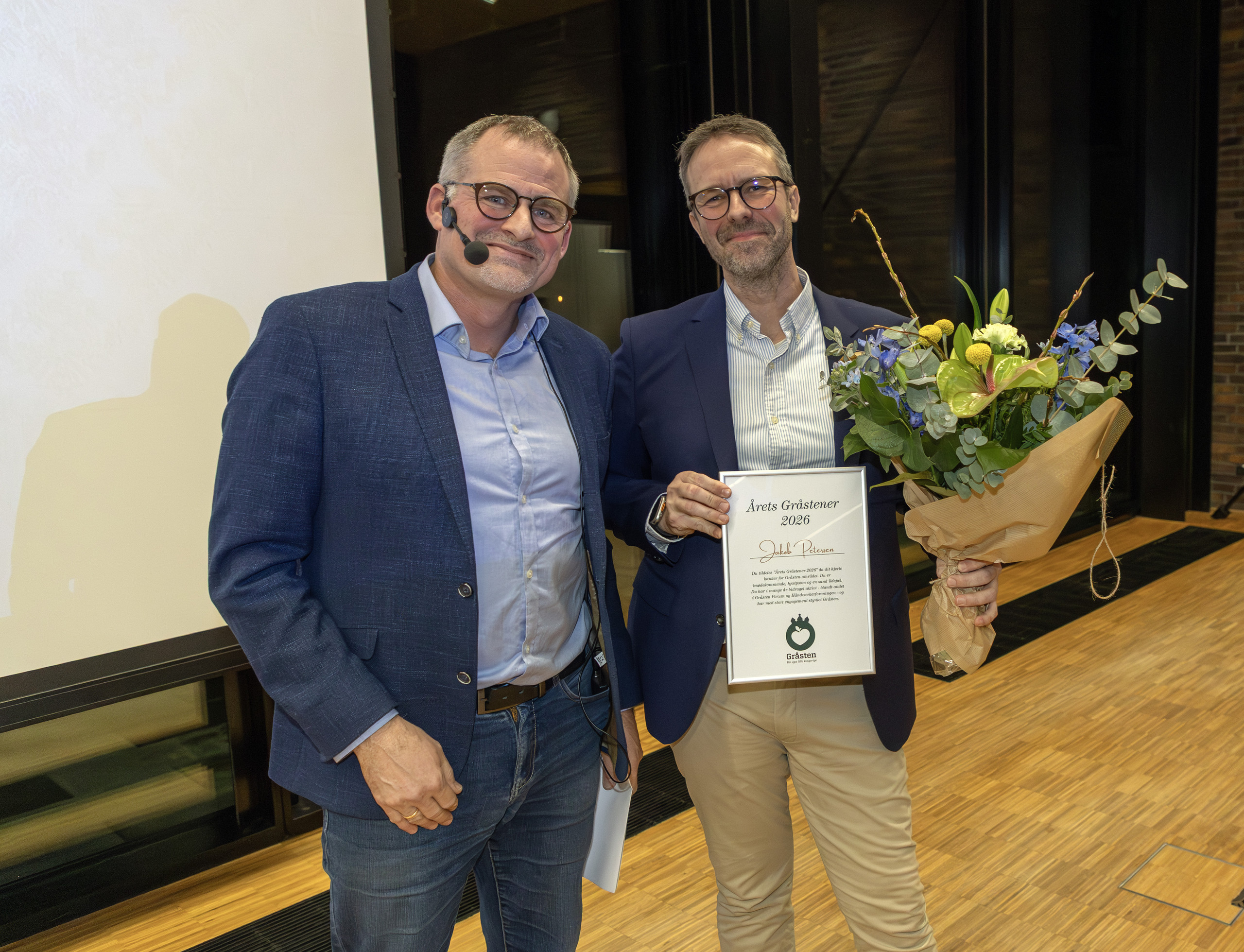 To mænd i mørke blazere står sammen på et trægulv, den ene holder et diplom og en buket blomster