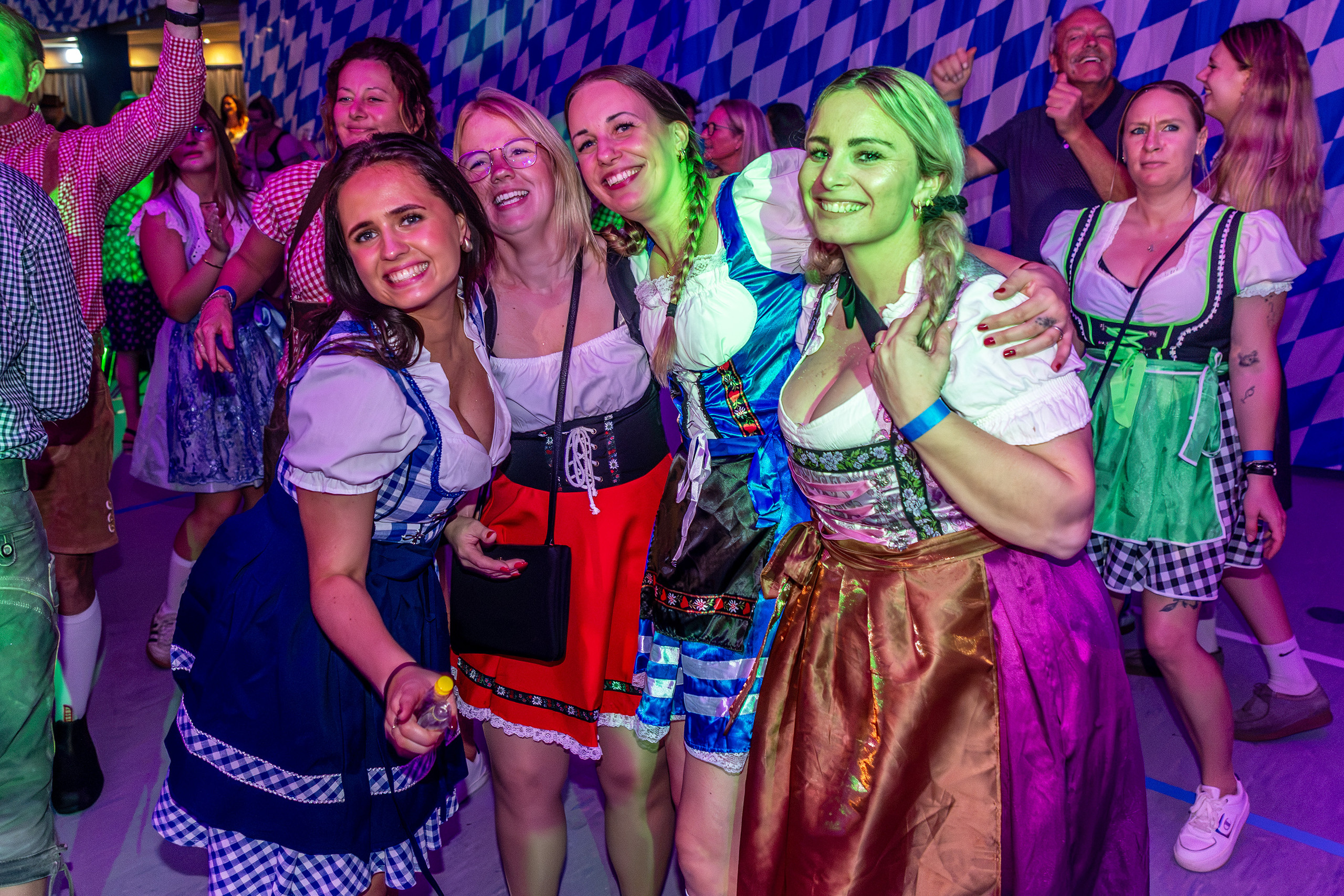Fem unge kvinder i farverige dirndl-kjoler poserer sammen med armene om hinanden