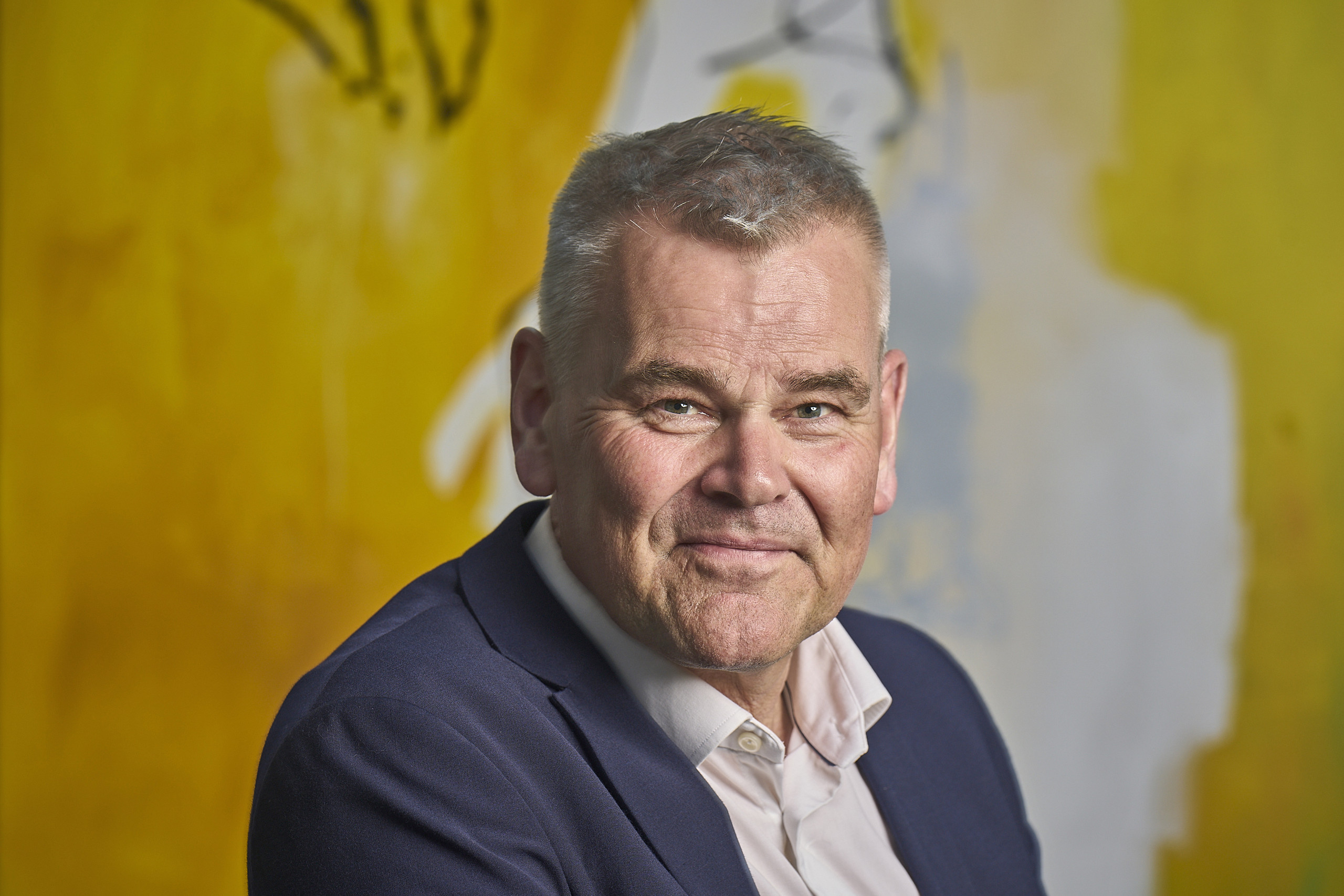 Portræt af smilende mand i mørkeblå jakke foran en gul væg