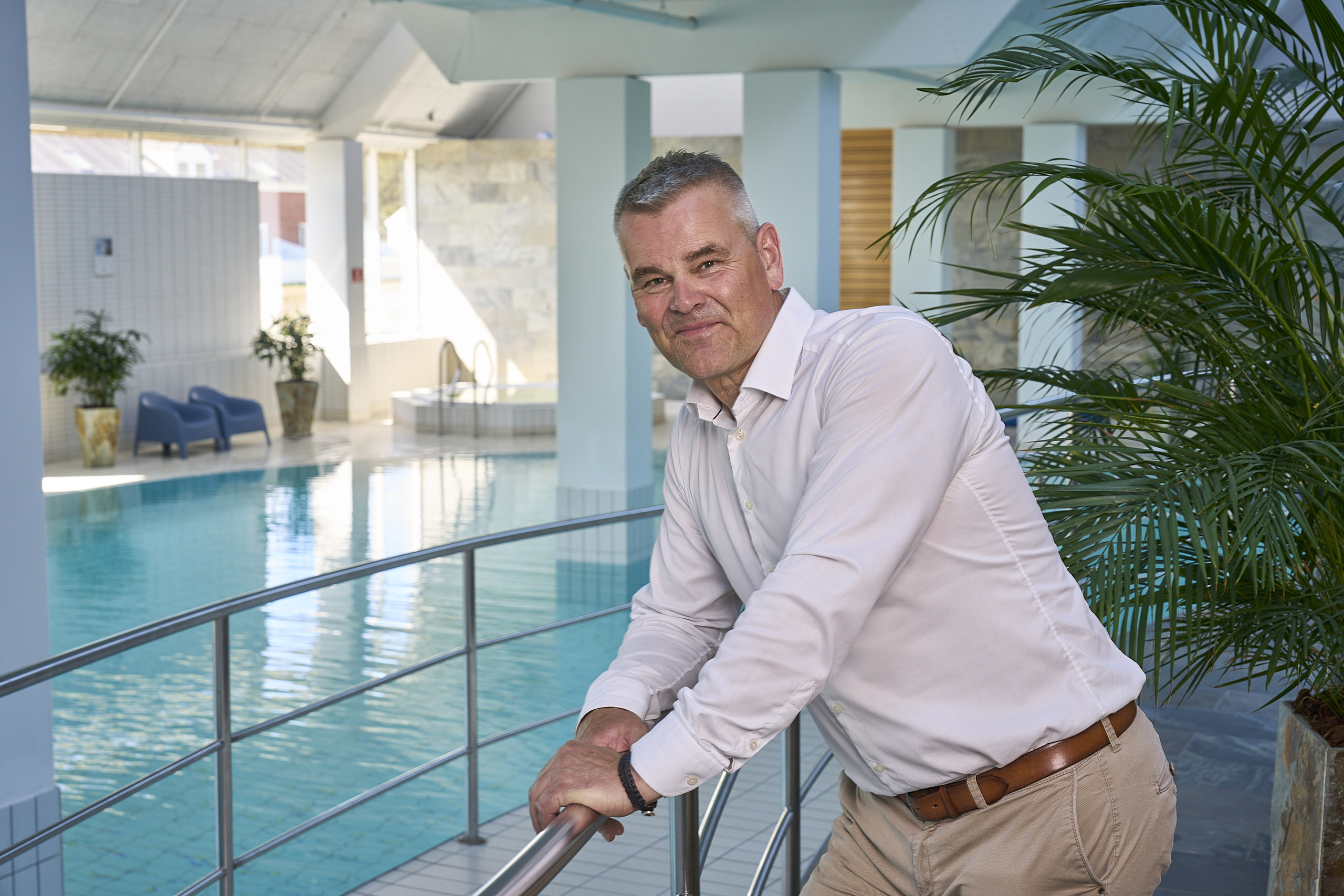 Mand i hvid skjorte læner sig op ad et gelænder ved en indendørs swimmingpool