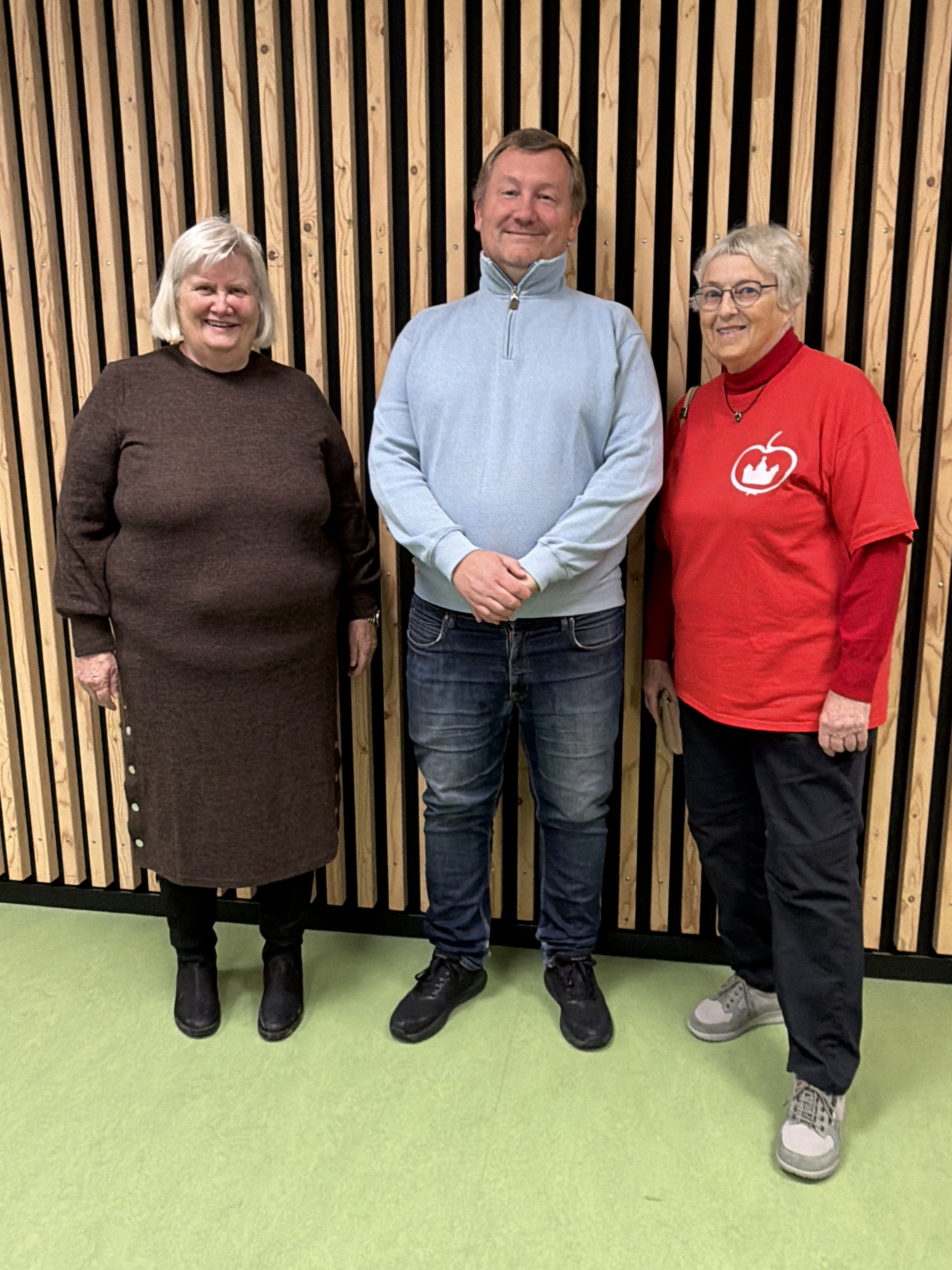 En mand i lyseblå trøje står mellem to kvinder foran en væg af lodrette trælister, den ene kvinde i rød trøje med æble-logo