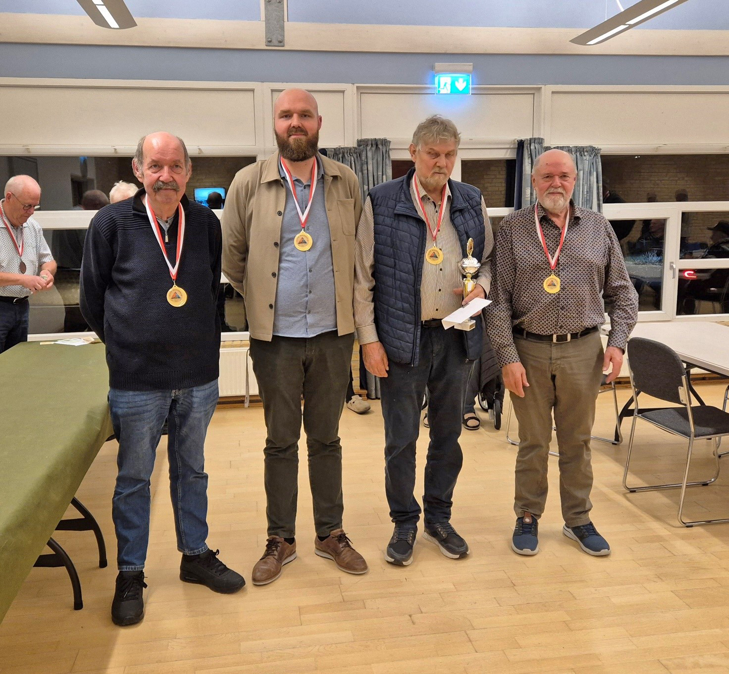 Fire mænd med medaljer om halsen står side om side i en sal, en af dem holder en pokal