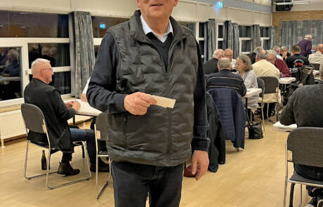 Mand i sort vest står med et stykke papir i hånden foran spillere ved bordene