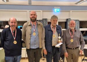 Fire mænd med medaljer om halsen står side om side i en sal, en af dem holder en pokal