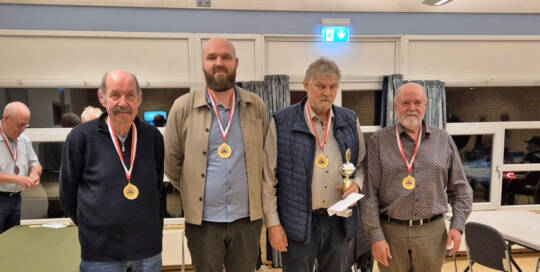 Fire mænd med medaljer om halsen står side om side i en sal, en af dem holder en pokal