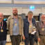 Fire mænd med medaljer om halsen står side om side i en sal, en af dem holder en pokal