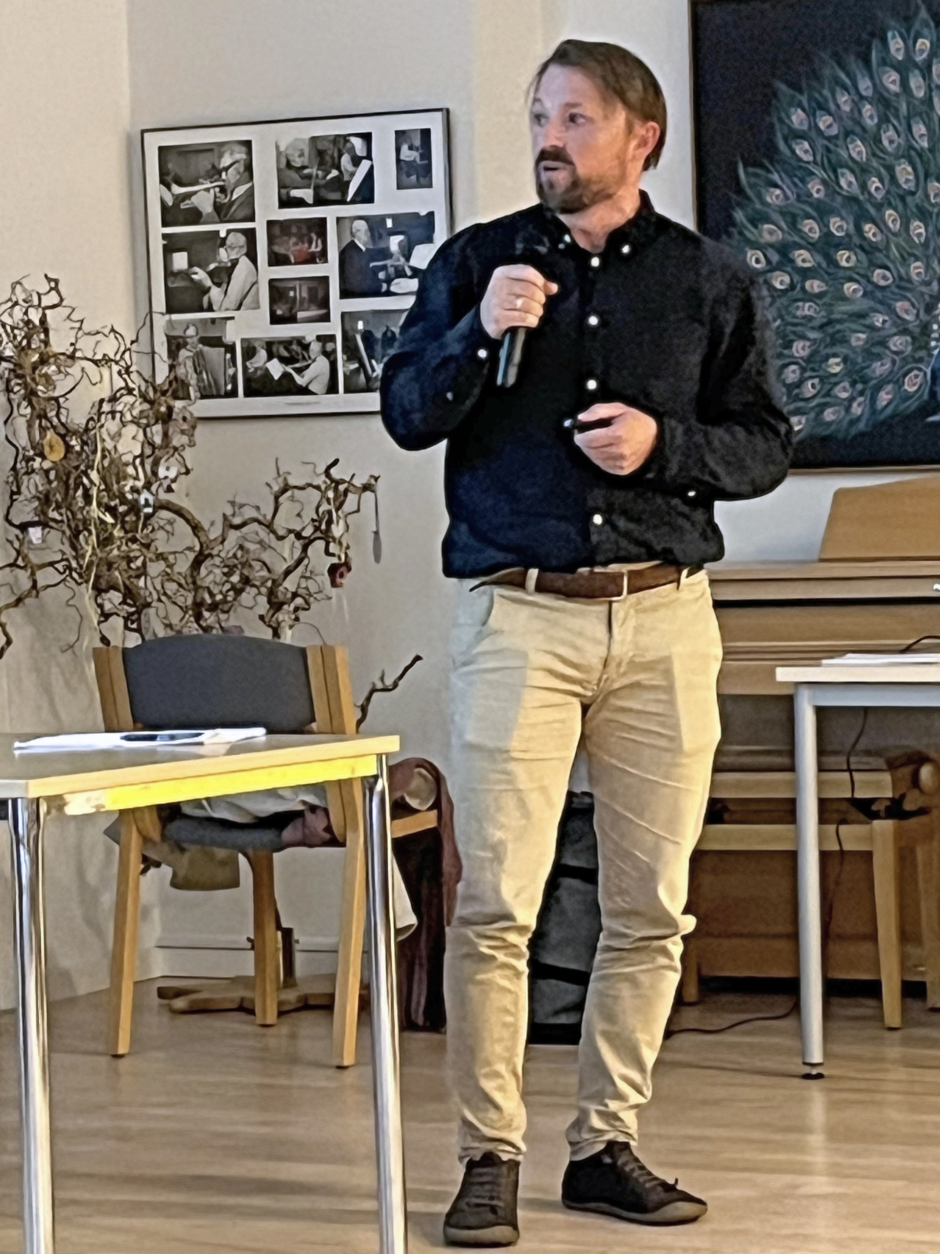 Mand i sort skjorte taler i mikrofon foran en opslagstavle med sort-hvide billeder