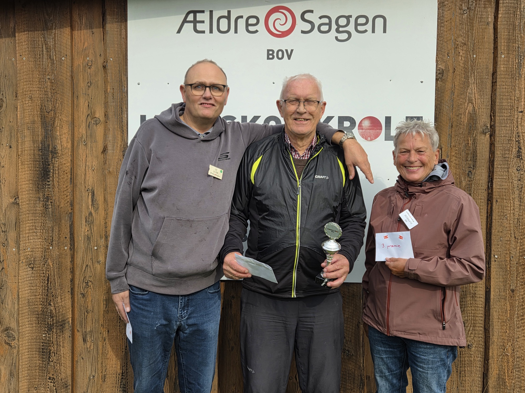 Tre personer står foran et skilt fra Ældre Sagen Bov, og en af dem holder en pokal