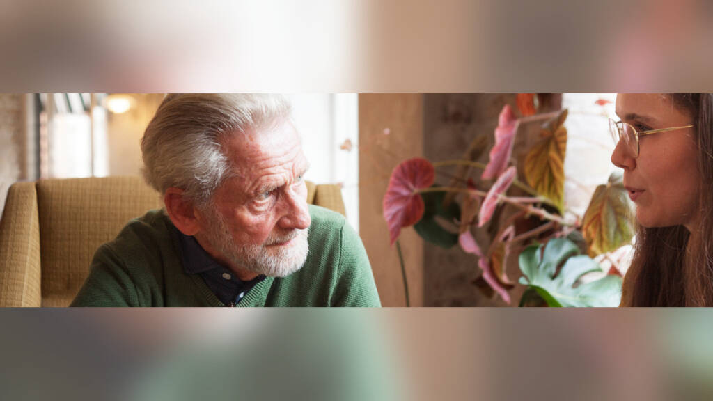 Ældre mand i grøn sweater sidder i en lænestol og taler med en ung kvinde