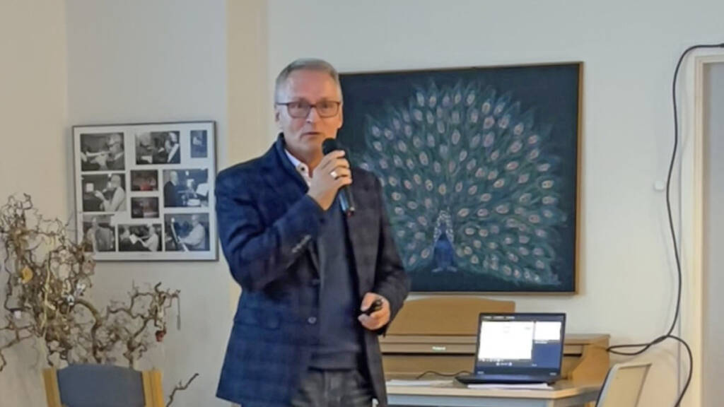 Mand i blå blazer taler i mikrofon i et lokale med et stort maleri af en påfugl på væggen