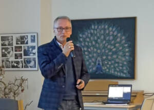Mand i blå blazer taler i mikrofon i et lokale med et stort maleri af en påfugl på væggen