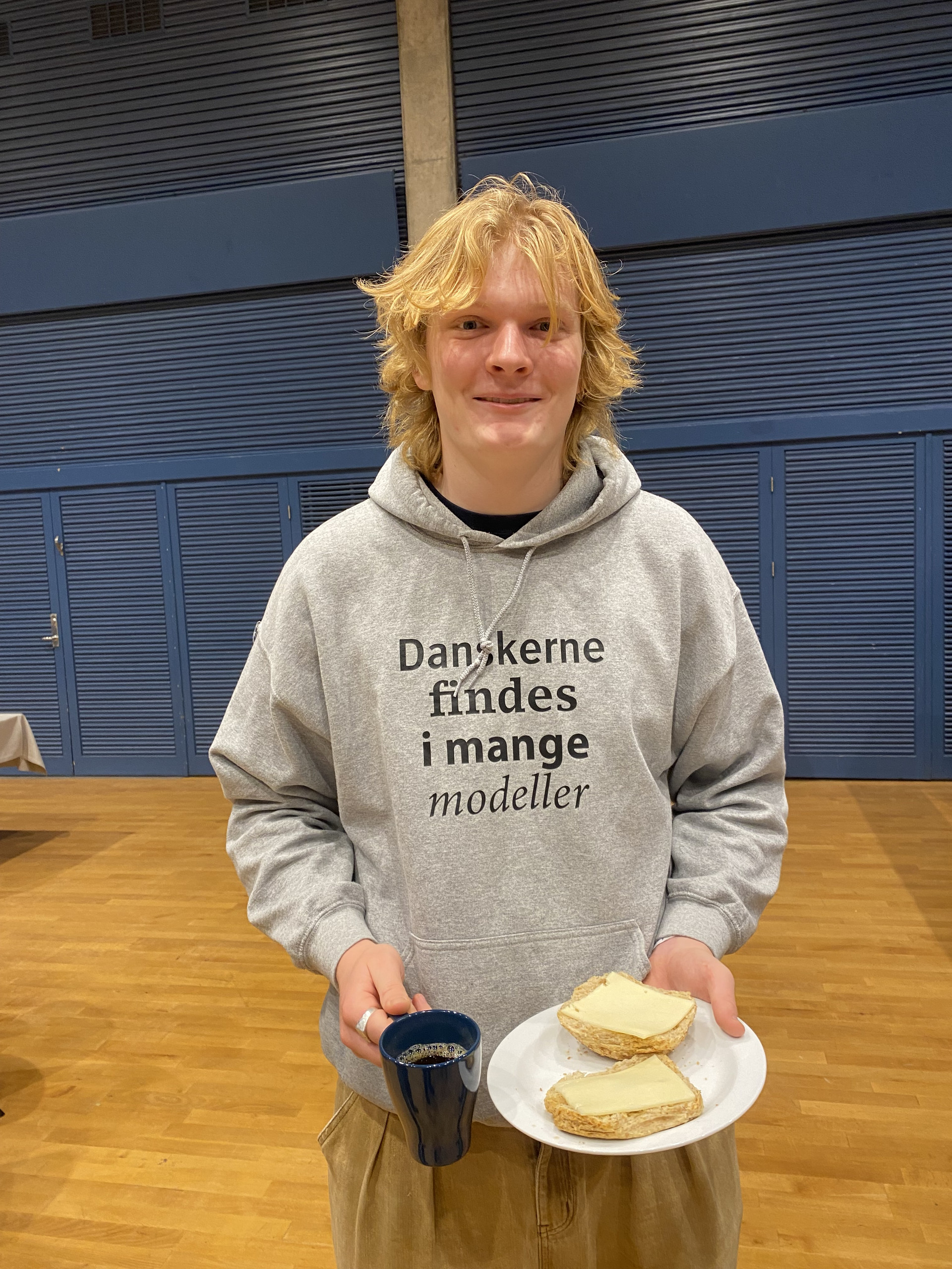 Ung mand i grå hættetrøje med teksten Danskerne findes i mange modeller holder en kop og en tallerken med to stykker brød i en hal