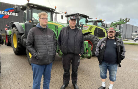 Tre mænd står foran grønne traktorer på en asfalteret plads