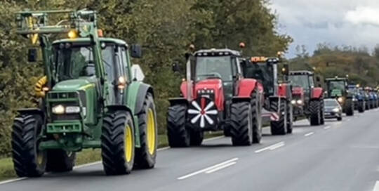 Lang række traktorer i konvoj på en landevej, forrest en grøn og en rød traktor