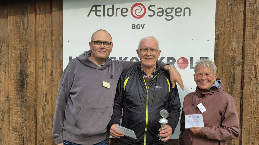 Tre personer står foran et skilt fra Ældre Sagen Bov, og en af dem holder en pokal