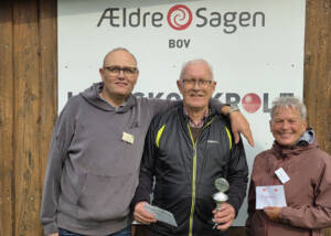 Tre personer står foran et skilt fra Ældre Sagen Bov, og en af dem holder en pokal